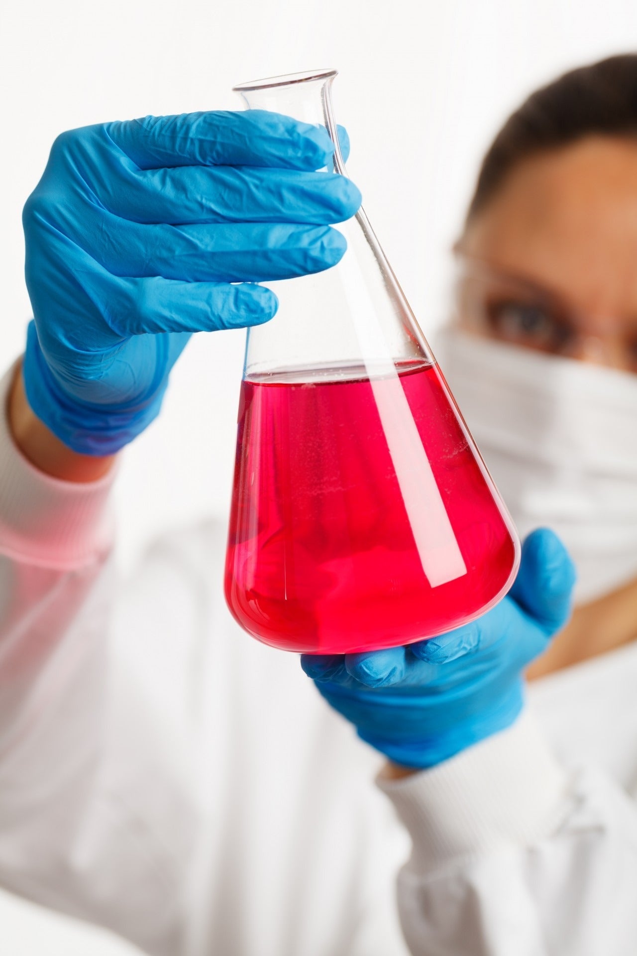 Woman dressed in white with a white mask looking at a laboratory glass with red liquid