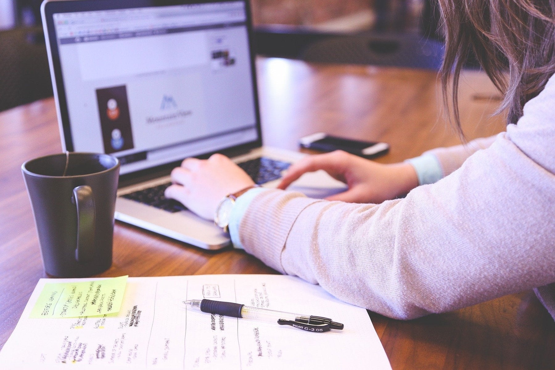 Woman working on a laptop surrounded by a black cup and a notebook