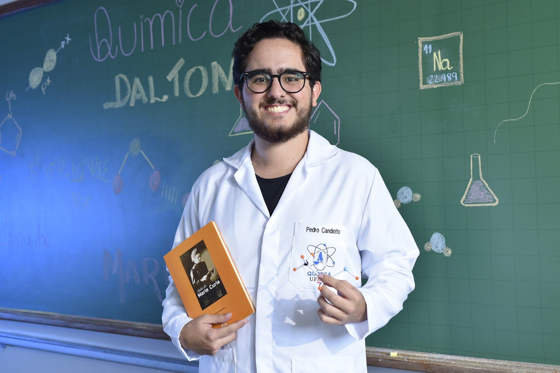 Smiling man dressed in white holding a book in front of a board