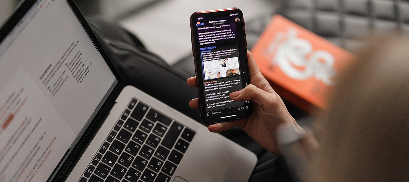 Woman holding phone and scrolling with a laptop beneath