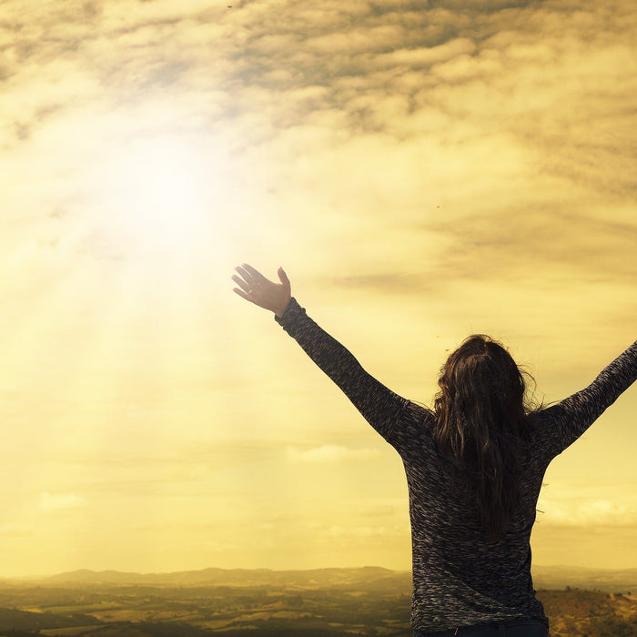 Woman with rised hand looking at the sun coming out of clouds over a field
